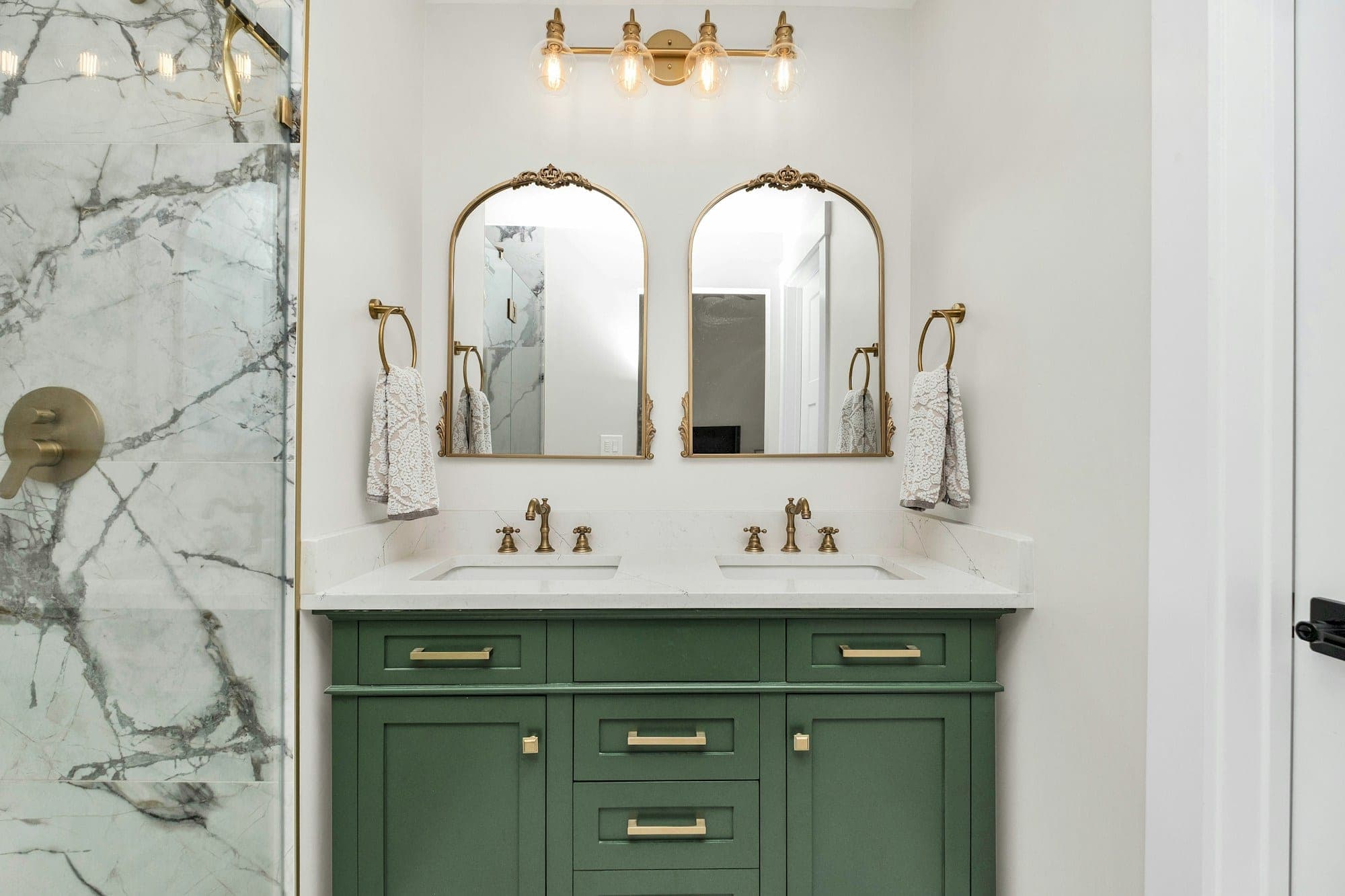 Bathroom with a green shaker vanity, marble top, arched mirrors and brass fixtures