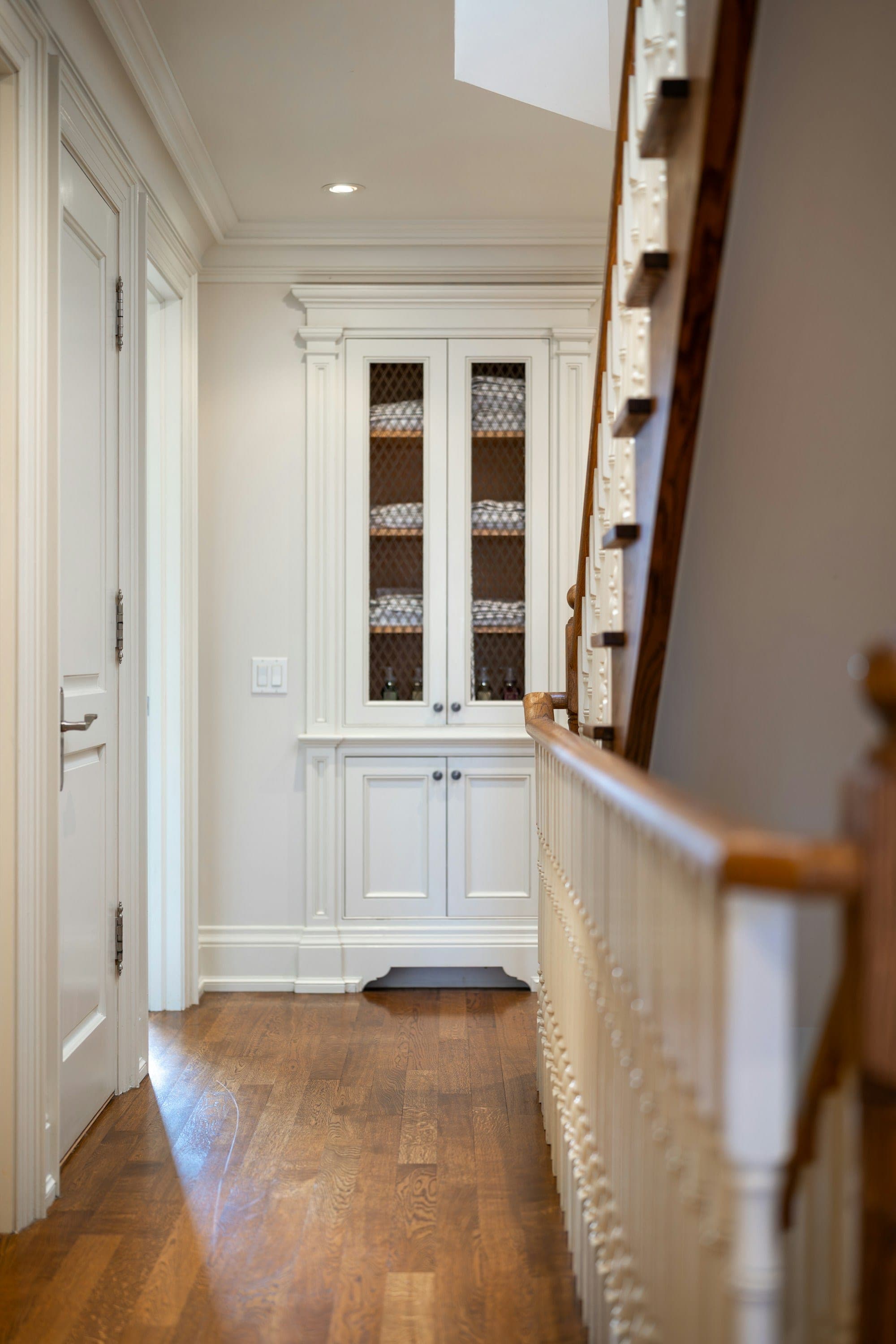 Painted built-in hallway cabinet with glass upper doors next to a wood stair railing